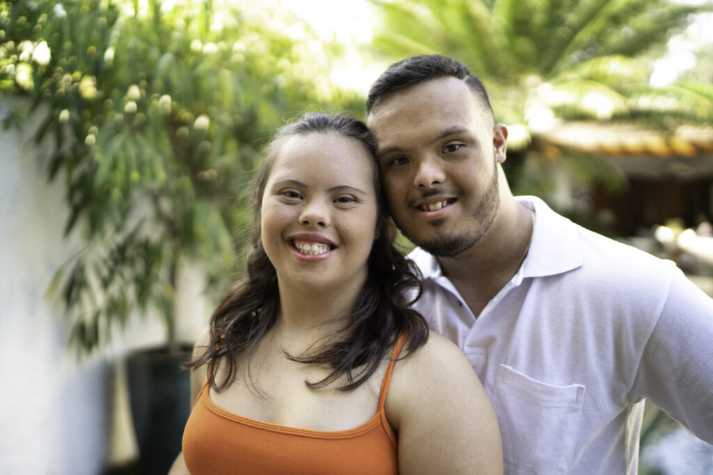 A young woman with Down syndrome smiles brightly, wearing an orange tank top, standing next to a young man with Down syndrome in a white shirt. They are outdoors, surrounded by greenery, with a warm, sunny atmosphere.