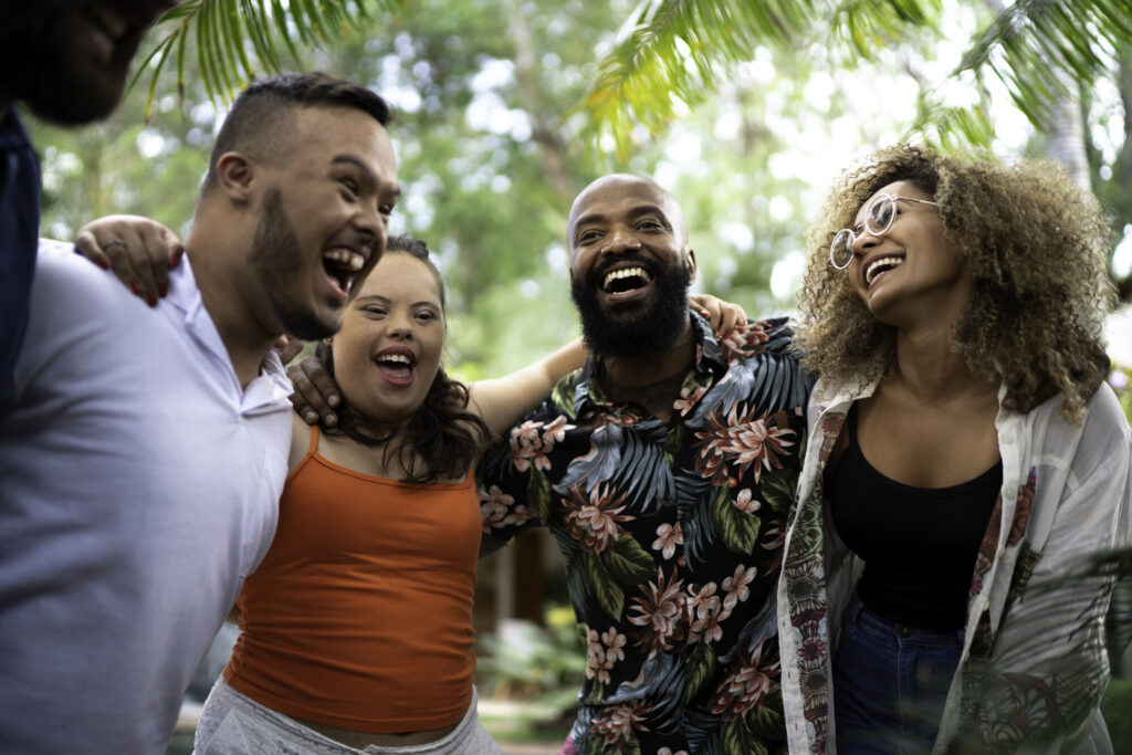 A diverse group of four friends stands together outdoors, laughing and enjoying each other's company. They are surrounded by greenery, with bright smiles and casual clothing, creating a joyful and inclusive atmosphere.