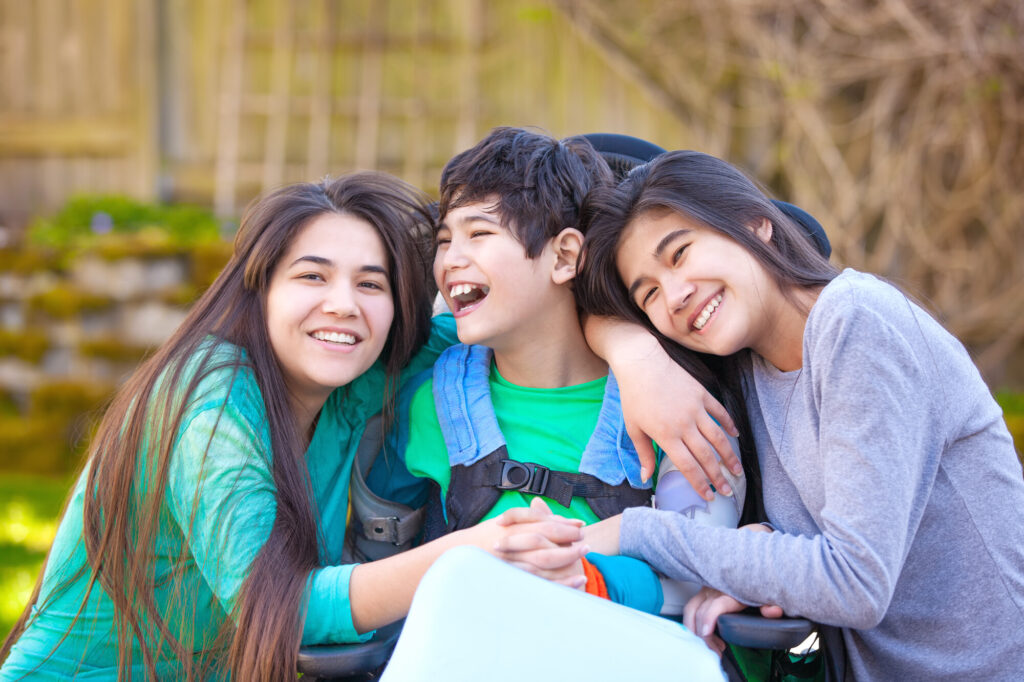 Three children are smiling and embracing each other outdoors. One child is in a wheelchair while the other two are on either side, showing joy and affection.