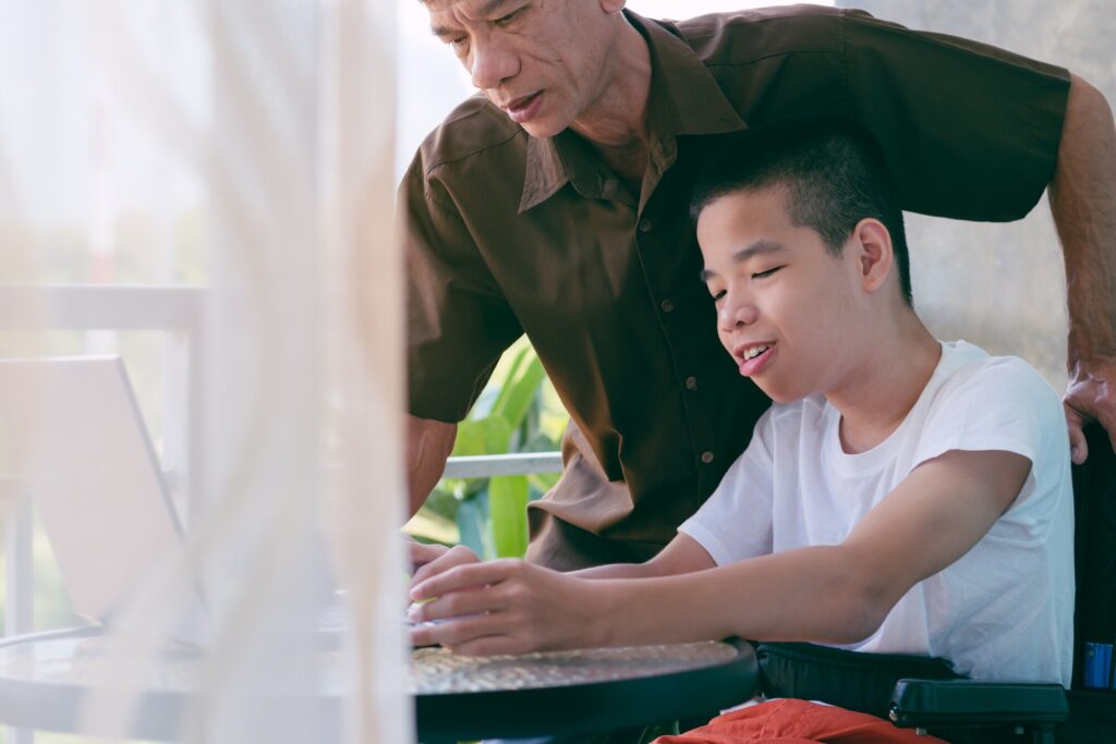 A young boy in a wheelchair smiles while interacting with a laptop, assisted by an older man. They are seated at a table, surrounded by greenery, with soft light filtering through a sheer curtain. The scene conveys a sense of connection and support.
