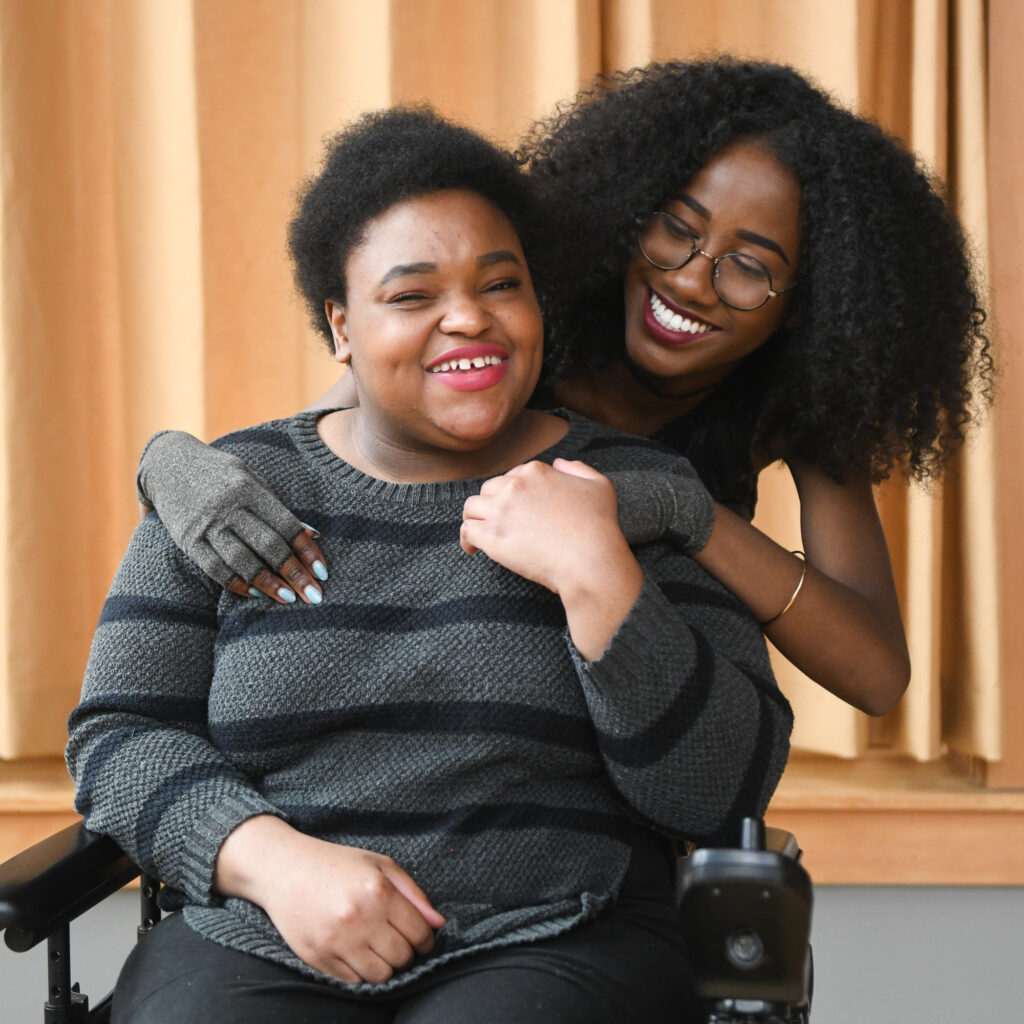 Two women are smiling together in a warm, indoor setting. One woman, in a wheelchair, wears a cozy striped sweater, while the other stands behind her, playfully embracing her. Both have natural hairstyles and are enjoying a joyful moment together.
