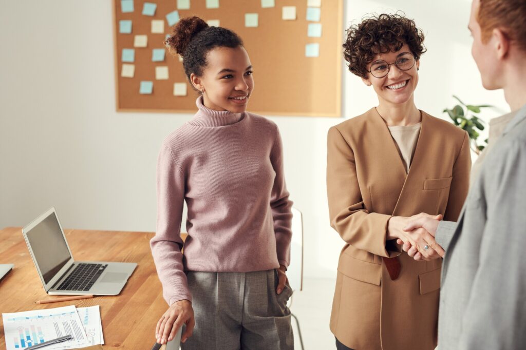 A professional setting featuring two women engaging in a handshake. One woman, wearing a beige blazer, smiles warmly, while the other, in a light pink sweater, stands nearby, looking pleased. A laptop and documents are visible on the table.