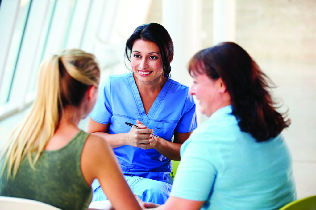 A healthcare professional in blue scrubs smiles while engaging in a conversation with two women. The setting is bright and modern, with large windows in the background, creating a welcoming atmosphere for discussion.