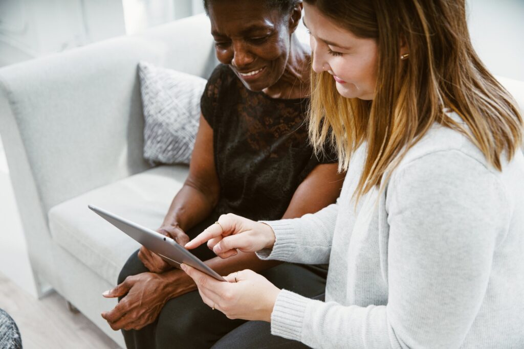A woman with long hair and a light sweater sits beside an older woman with short hair, both smiling as they look at a tablet together. They are seated on a light-colored couch in a bright, modern room.