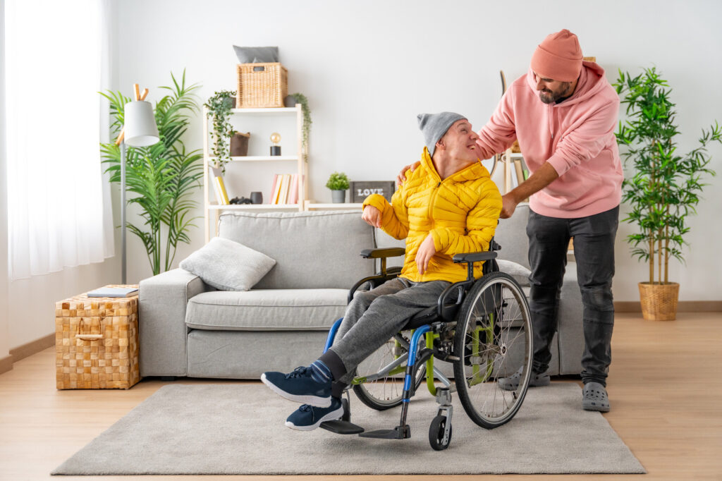 A young man in a yellow jacket sits in a wheelchair, smiling at another man who is standing beside him, gently placing a hand on his shoulder. They are in a bright, modern living room with plants and a cozy sofa