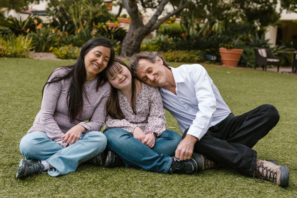 A smiling family of three sits on green grass in a park. The mother and father lean in close to their daughter, who is in the middle. They are all casually dressed, enjoying a sunny day surrounded by trees and plants.