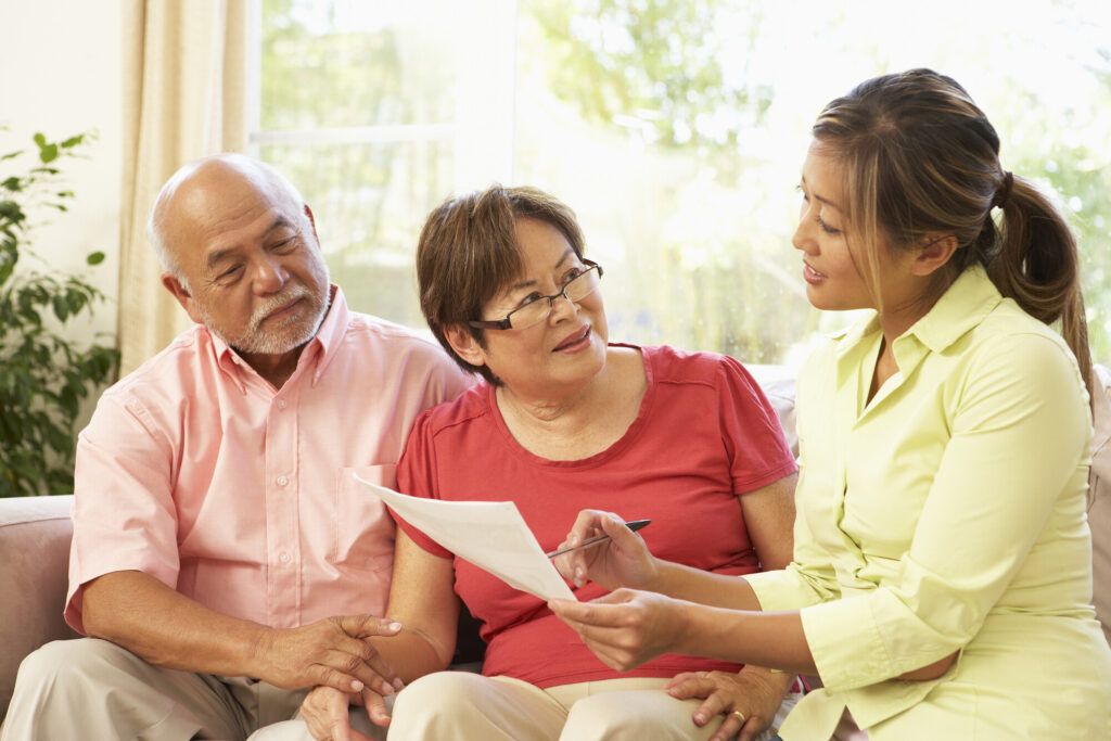 A young woman in a light green shirt is sitting with an older couple on a couch. She is holding a document and explaining something to them. The couple appears engaged and attentive, with a bright, sunny background visible through a window.