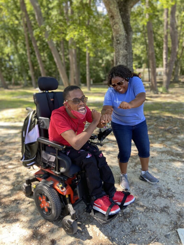 A person in a red shirt and wheelchair smiles while interacting with a woman in a blue shirt. They are outdoors, surrounded by trees, sharing a joyful moment together. The scene conveys friendship and support.