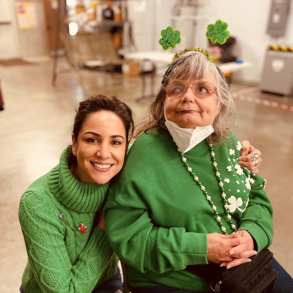 A woman in a green sweater smiles while sitting next to an older woman wearing a green outfit and festive shamrock accessories. They are indoors, with a warm atmosphere, celebrating a festive occasion.