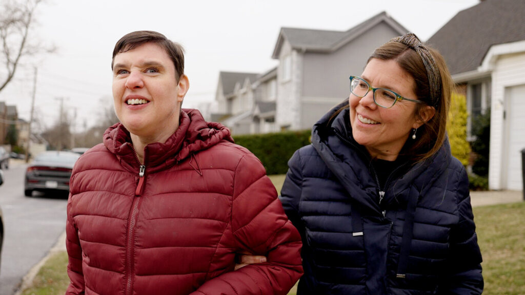 Two women are walking together on a suburban street. One woman, wearing a red jacket, is smiling and looking at the other, who is wearing a dark blue jacket and glasses. They appear to be enjoying each other's company on a cloudy day.